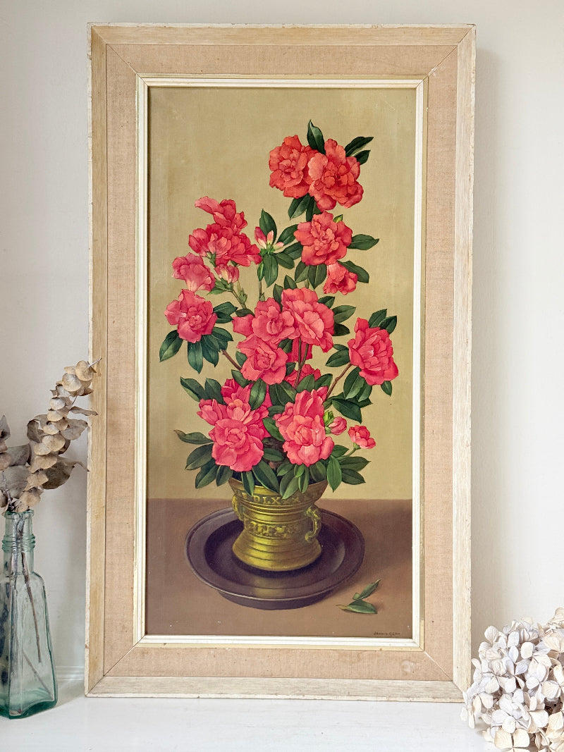 A framed vintage-style floral painting featuring bright pink-red peonies with green leaves in a gold vase, displayed against a light wall with decorative dried flowers and a glass bottle at the sides.