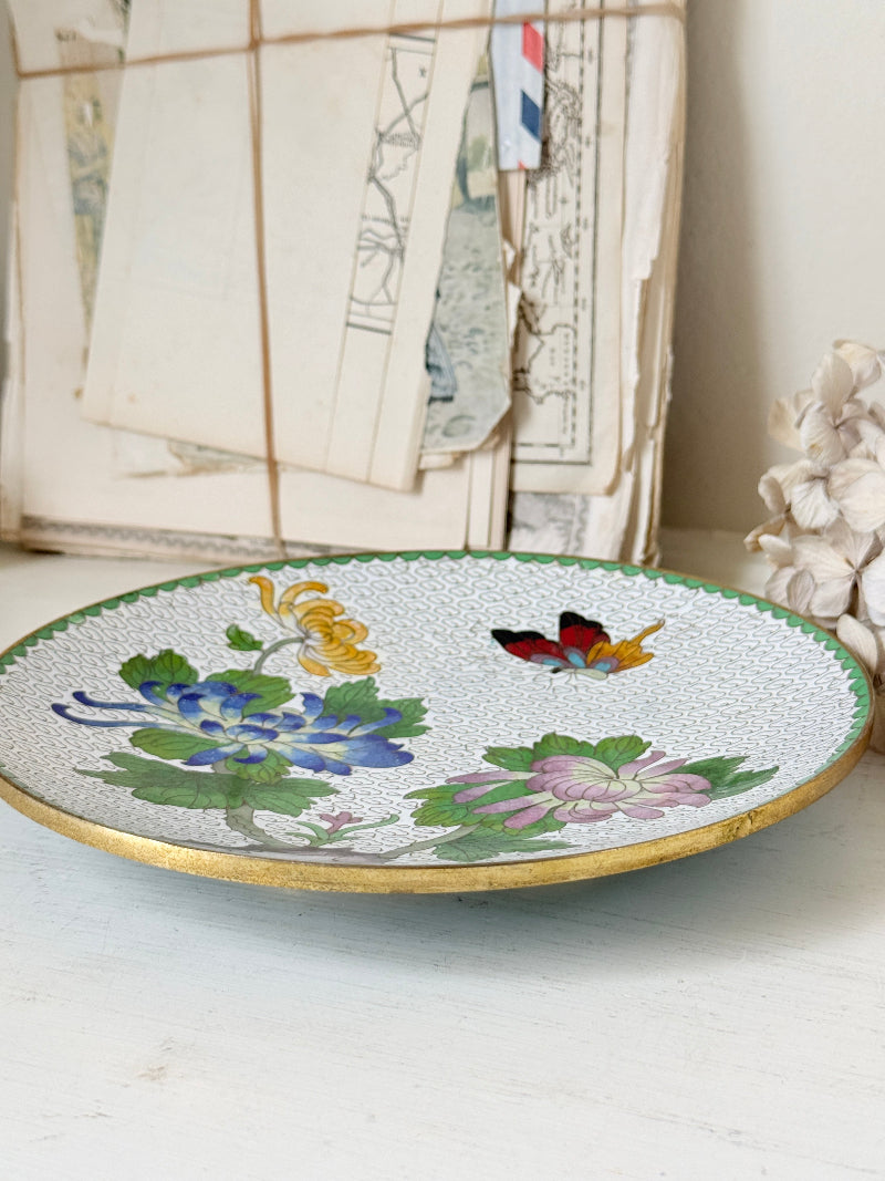 A round ceramic plate with a textured white glaze, decorated with colorful hand-painted flowers, butterflies, and a gold rim, styled on a white surface with vintage papers in the background.