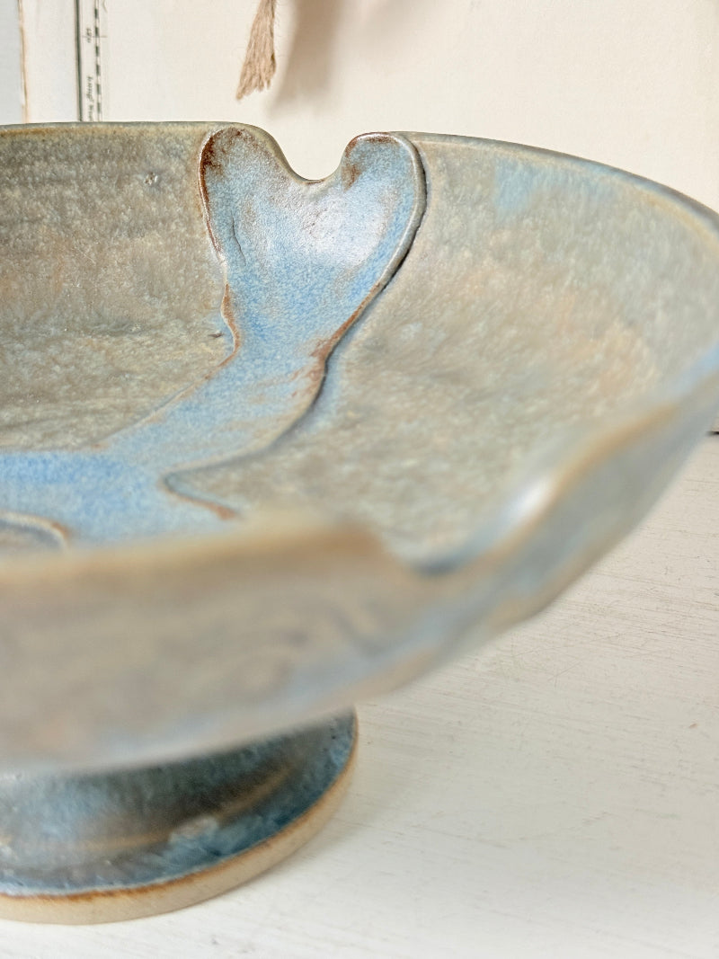 Close-up of a ceramic pedestal bowl with a textured glaze in muted blue, beige, and gray tones, set against a soft neutral background.