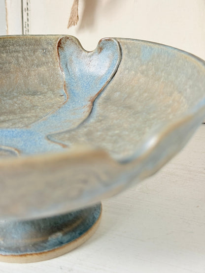 Close-up of a ceramic pedestal bowl with a textured glaze in muted blue, beige, and gray tones, set against a soft neutral background.