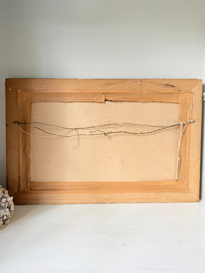 The back of a square wooden picture frame with a light natural wood finish, showing the backing board and hanging wire, resting on a white surface against a white wall.