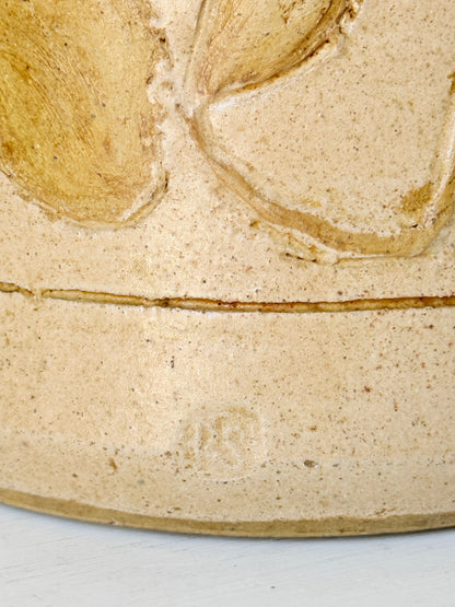 Close-up of a beige ceramic surface with a raised floral relief and a horizontal seam, showing a speckled clay texture and a faint stamped mark near the bottom.