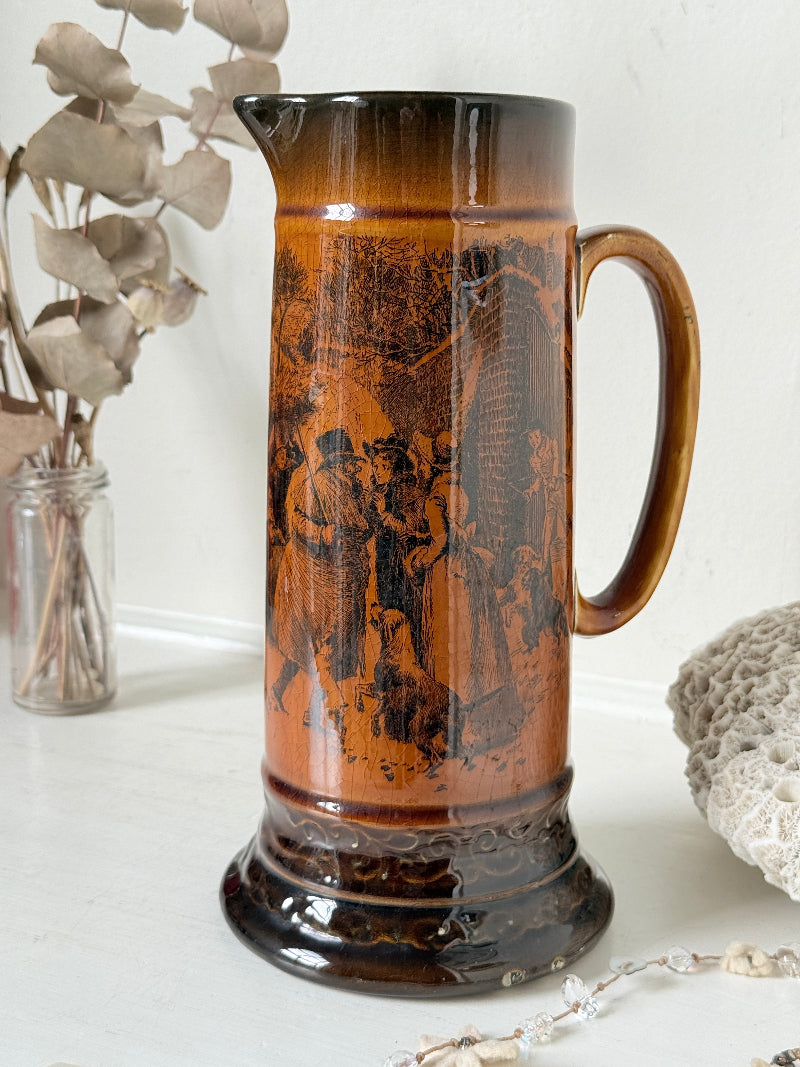 Antique Ridgways stoneware jug photographed on a white surface in amber treacle glaze with A Christmas Visitor black transfer scene and tall handle