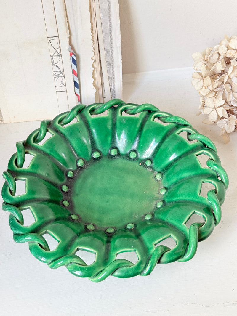 A green ceramic decorative bowl with a wavy, scalloped rim and cutout details, displayed on a white surface with neutral background decor.