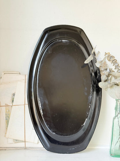 A large oval black serving platter or tray with a glossy finish, leaning against a white wall next to a green glass vase with dried white flowers.