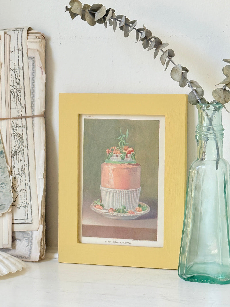 Framed Victorian cookery illustration of a celebration cake from 1896 in a mustard yellow frame displayed on a white surface.