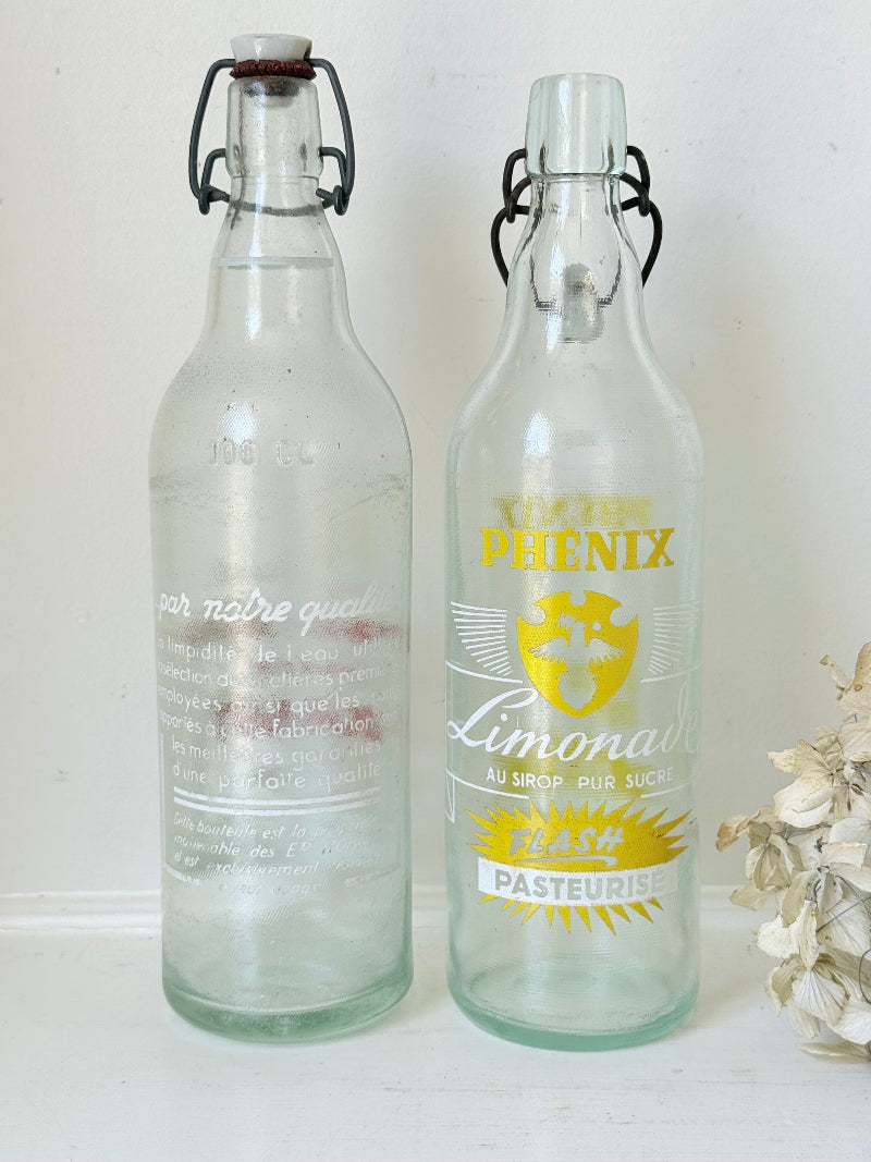 Pair of vintage French glass bottles with retro typography and graphics, photographed on a white surface