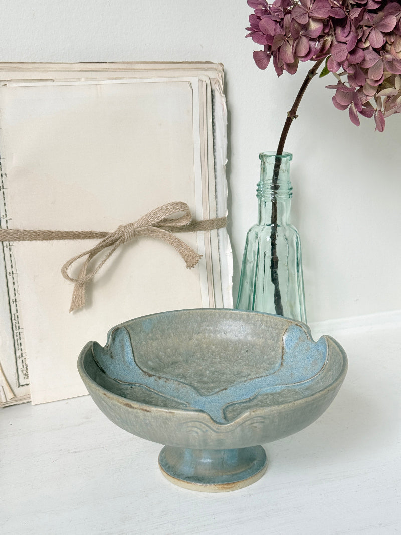 A shallow ceramic bowl with a pedestal base in a distressed light blue-gray glaze, styled on a white surface with a vintage-style cream box, a small clear glass bottle, and purple flowers in the background.