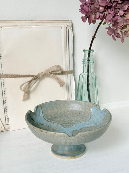 A shallow ceramic bowl with a pedestal base in a distressed light blue-gray glaze, styled on a white surface with a vintage-style cream box, a small clear glass bottle, and purple flowers in the background.