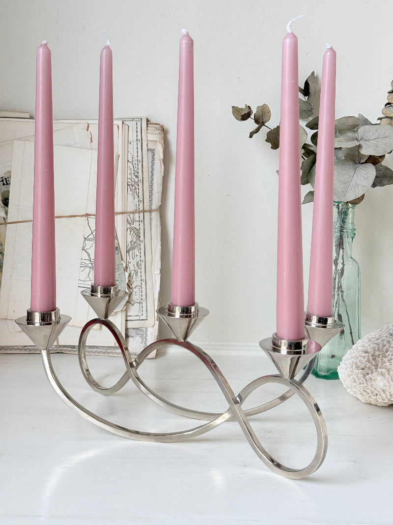 Silver tone five light candelabra with a sculptural looped base holding pink candles, photographed on a white surface