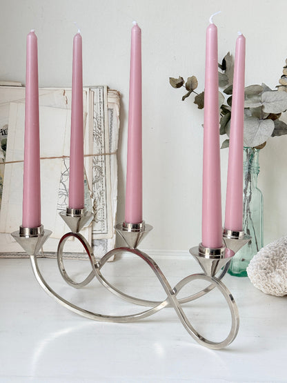 Silver tone five light candelabra with a sculptural looped base holding pink candles, photographed on a white surface