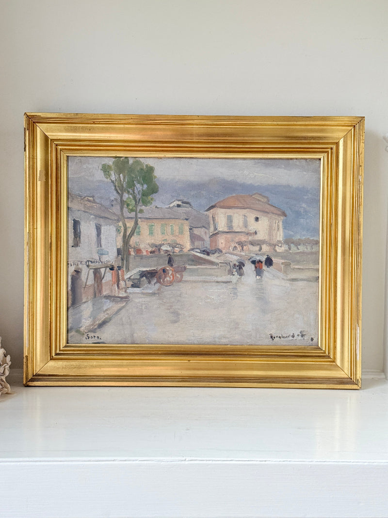 A framed impressionist landscape painting depicting a wet street scene with buildings, a tree, and figures, housed in a wide ornate gold frame and displayed on a white shelf against a light wall.