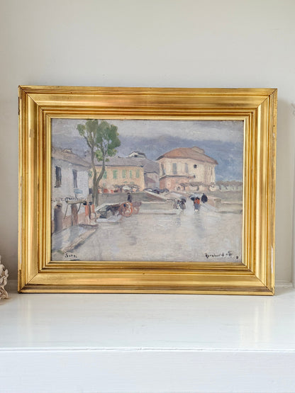 A framed impressionist landscape painting depicting a wet street scene with buildings, a tree, and figures, housed in a wide ornate gold frame and displayed on a white shelf against a light wall.