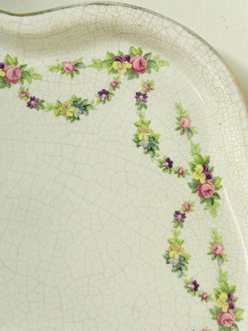 Vintage cream ceramic serving plate with scalloped wavy edge and pink and green floral garland border photographed on a white surface