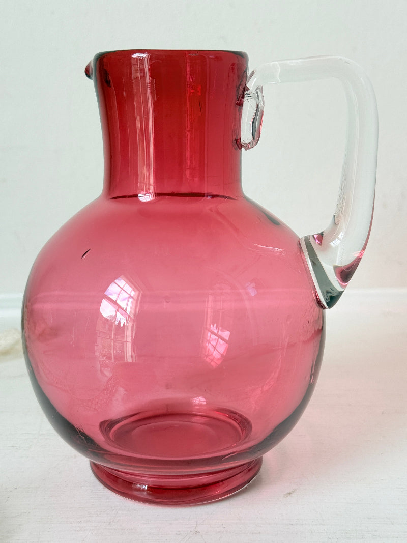 Vintage cranberry glass jug with clear handle photographed on a white surface, bulbous pink body with flared pouring rim