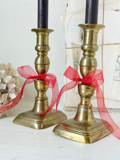 Pair of turned brass candlesticks with square stepped bases and black taper candles on a white surface, styled with dried flowers