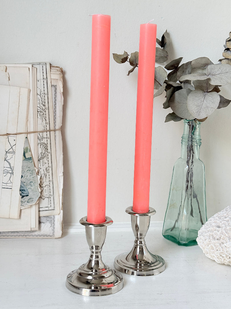 Pair of silver tone candlesticks with urn sconces and round stepped bases holding coral candles, photographed on a white surface