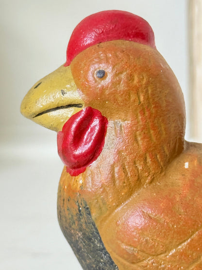 A close-up of a hand-painted wooden rooster figurine, showing its textured orange-brown body, bright red comb and wattle, and yellow beak.