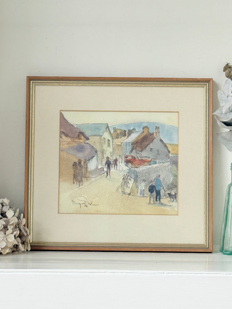 A framed watercolor painting of a village street scene with people walking, displayed on a white mantelpiece against a light wall.