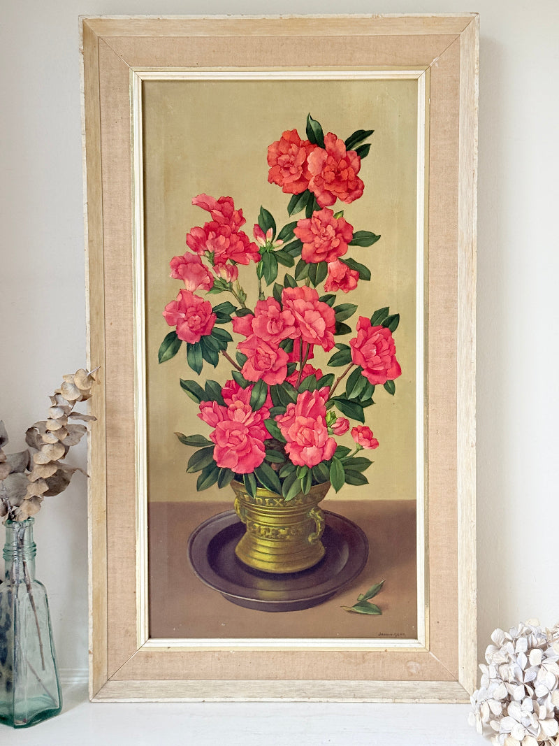 A framed vintage-style floral painting featuring bright pink flowers in a gold vase against a warm beige background, displayed on a shelf with dried botanicals and a glass bottle.