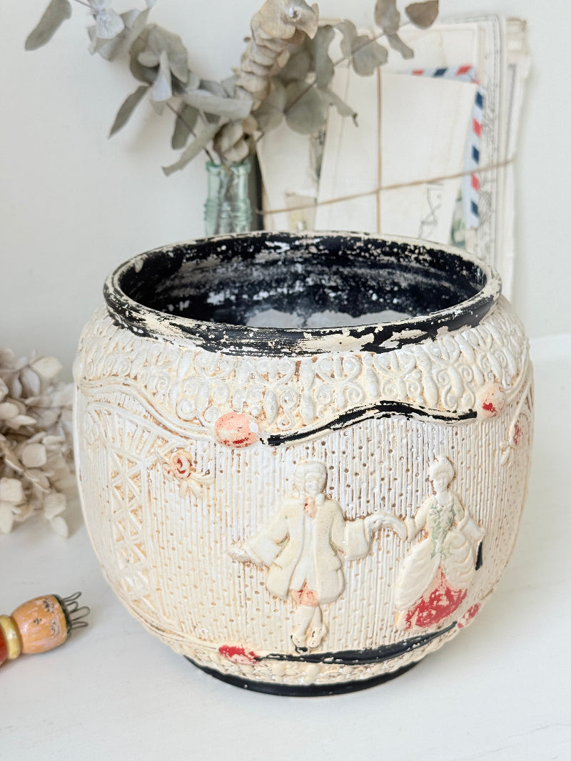 A round ceramic planter pot with a distressed cream-colored exterior featuring embossed figures and floral details, a black interior, and small pink and red accents, set against a softly blurred background with dried flowers.