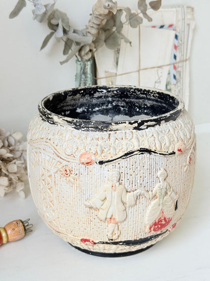 A round ceramic planter pot with a distressed cream-colored exterior featuring embossed figures and floral details, a black interior, and small pink and red accents, set against a softly blurred background with dried flowers.