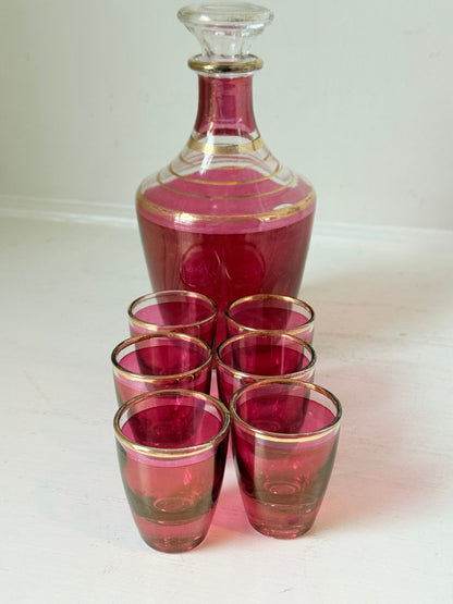 Vintage red printed glass cocktail set with clear glass decanter and six small glasses with gilt band rims, photographed on a white surface