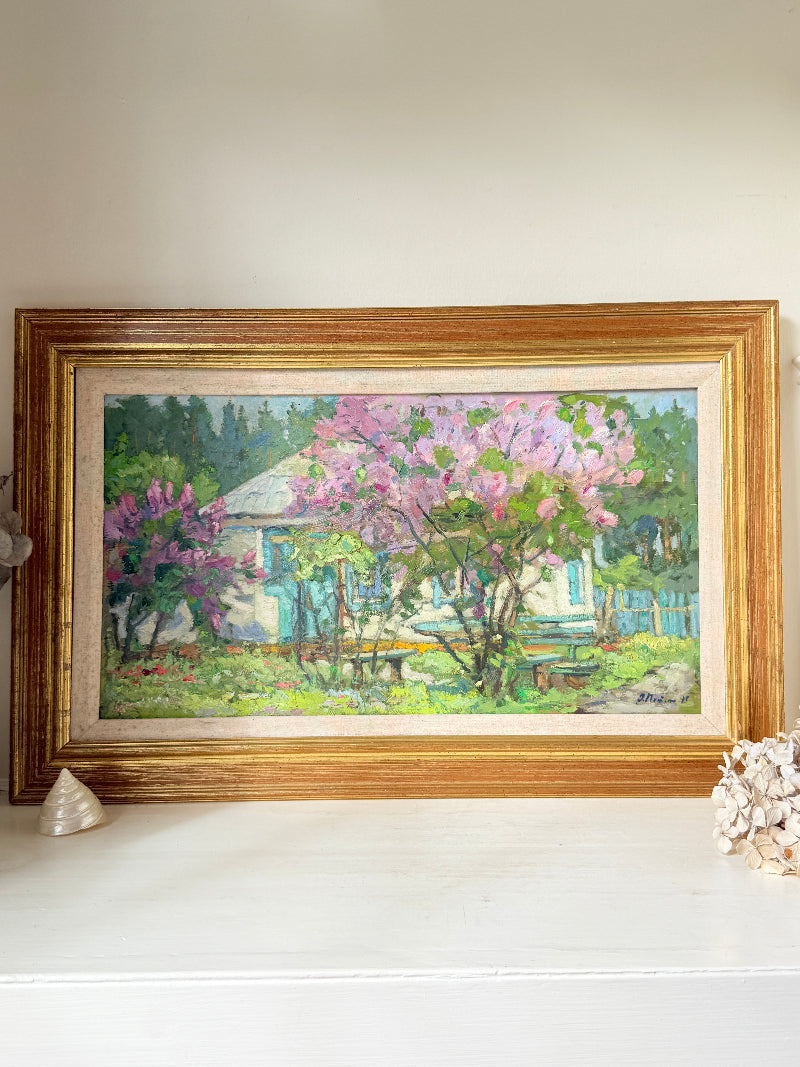 A framed oil painting of a cottage garden with pink flowering trees, displayed on a white shelf against a beige wall.