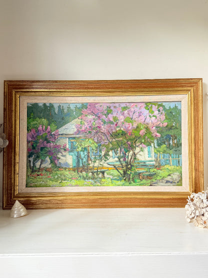 A framed oil painting of a cottage garden with pink flowering trees, displayed on a white shelf against a beige wall.