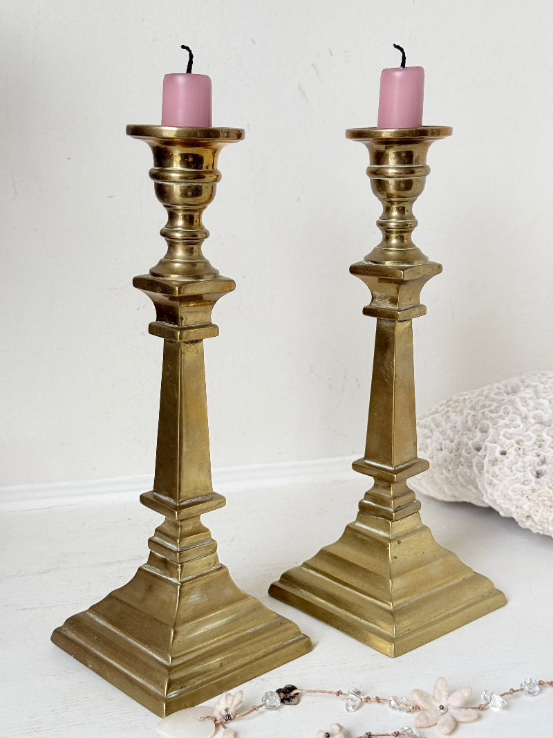 Pair of Georgian style brass candlesticks photographed on a white surface with tall tapered stems and stepped square bases holding pink taper candles