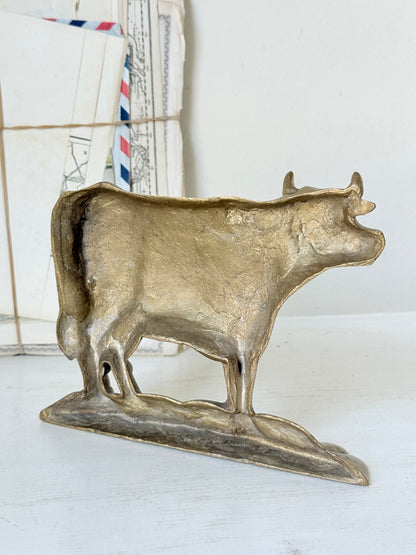 A rustic, hand-painted metallic cow figurine in a distressed gold/bronze finish, standing on a matching base against a white background with vintage mail in the soft-focus background.