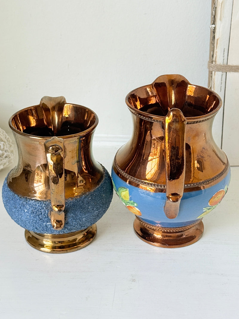 Two vintage-style metal pitchers with copper-toned bodies and blue lower sections, one plain and one with a small floral pattern, displayed on a white surface against a white wall.