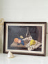 A framed still life oil painting depicting fruit, grapes, a pear, and a glass bottle on a tablecloth, set in a dark brown wooden frame with a cream mat, leaning against a light wall.