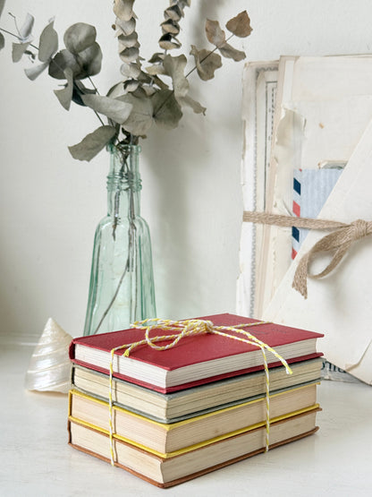 A stack of four vintage hardcover books tied with a yellow string, set on a white surface next to a glass bottle vase with dried eucalyptus and a rustic framed print.