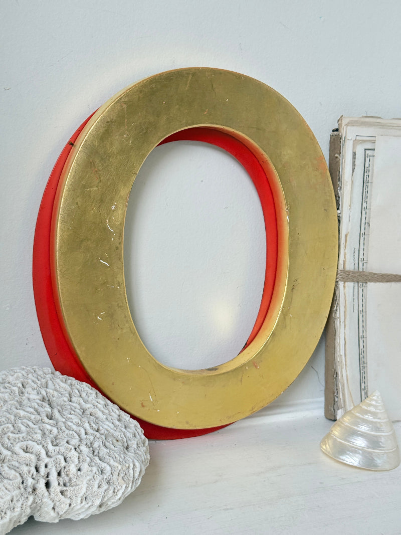 A large decorative letter O wall sign with a distressed gold outer ring and a red inner ring, styled on a white surface with a white coral stone and a clear seashell.