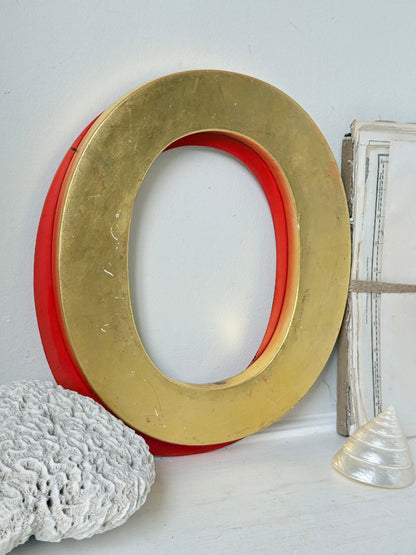 A large decorative letter O wall sign with a distressed gold outer ring and a red inner ring, styled on a white surface with a white coral stone and a clear seashell.