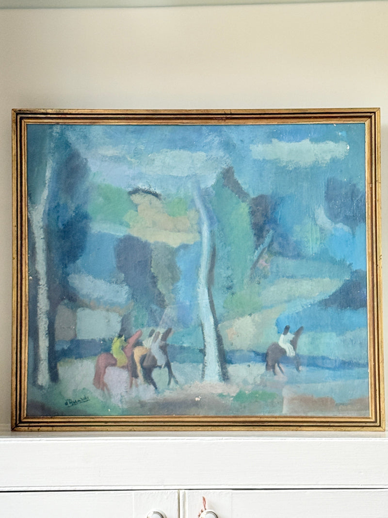 An impressionist-style landscape painting in a gold frame, hanging on a beige wall above a white cabinet. The painting depicts figures on horseback in a blue-toned forest scene.