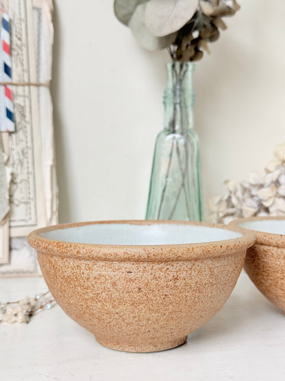 A round ceramic bowl with a speckled tan exterior and a pale blue interior, shown on a tabletop with a matching bowl partially visible at the right and decorative objects in the background.
