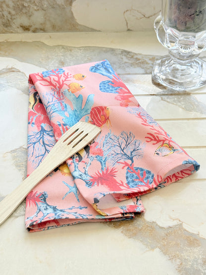 Warm peach coral and tropical fish print cotton tea towel with wooden forks on a marble surface beside cut glass