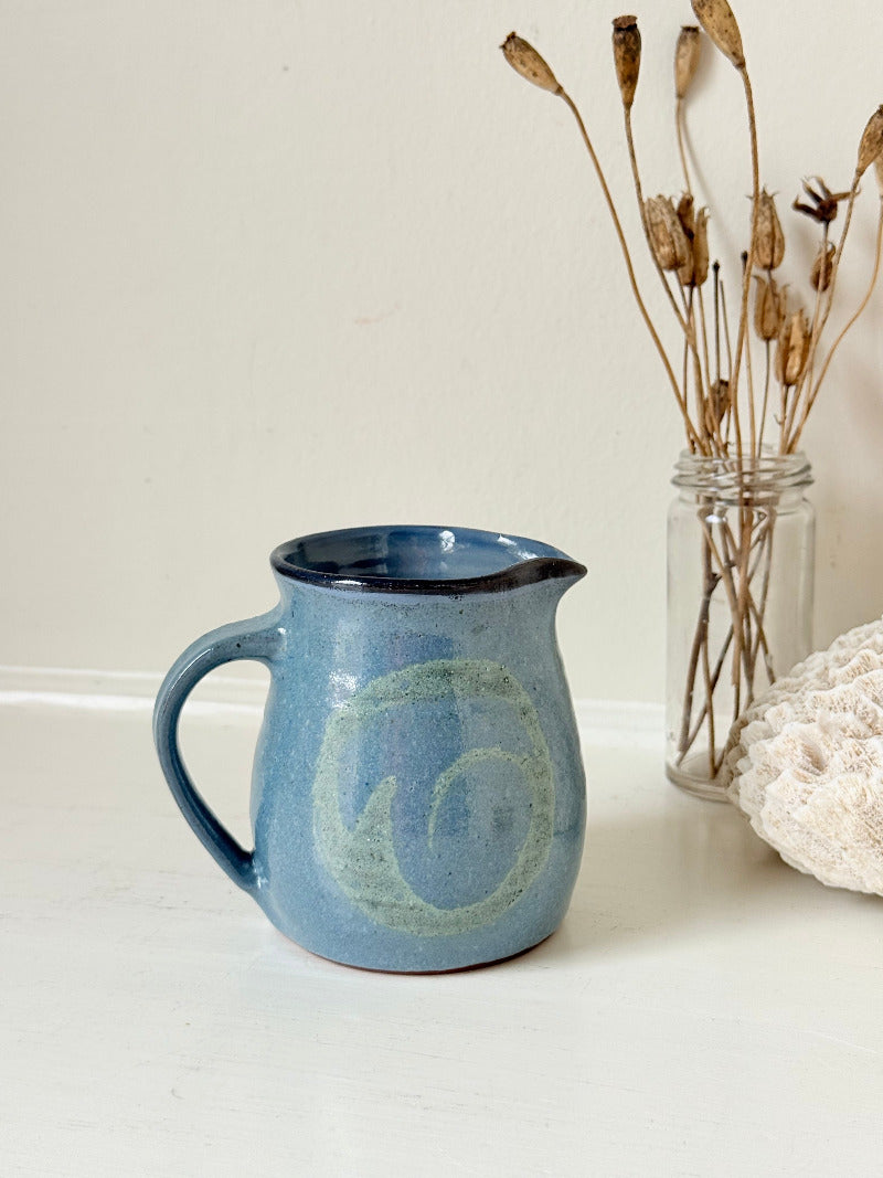 Vintage studio pottery milk jug with blue and green glaze, handmade with a rounded form and sturdy handle, styled on a white surface.
