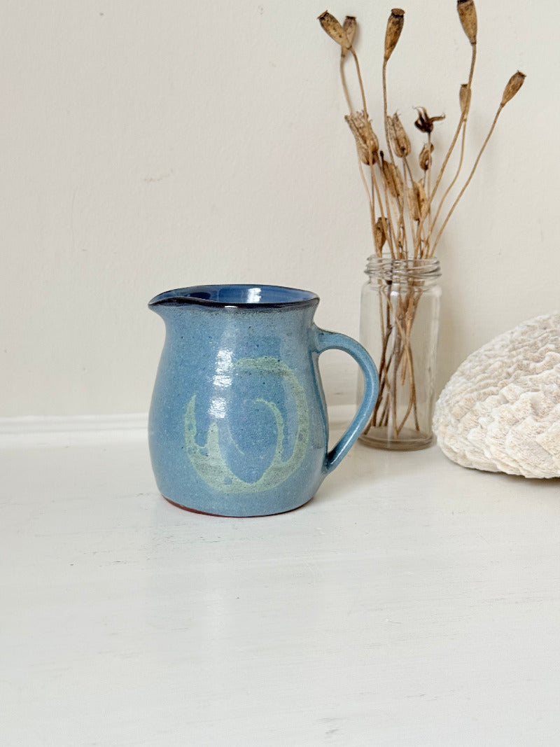 Vintage studio pottery milk jug with blue and green glaze, handmade with a rounded form and sturdy handle, styled on a white surface.