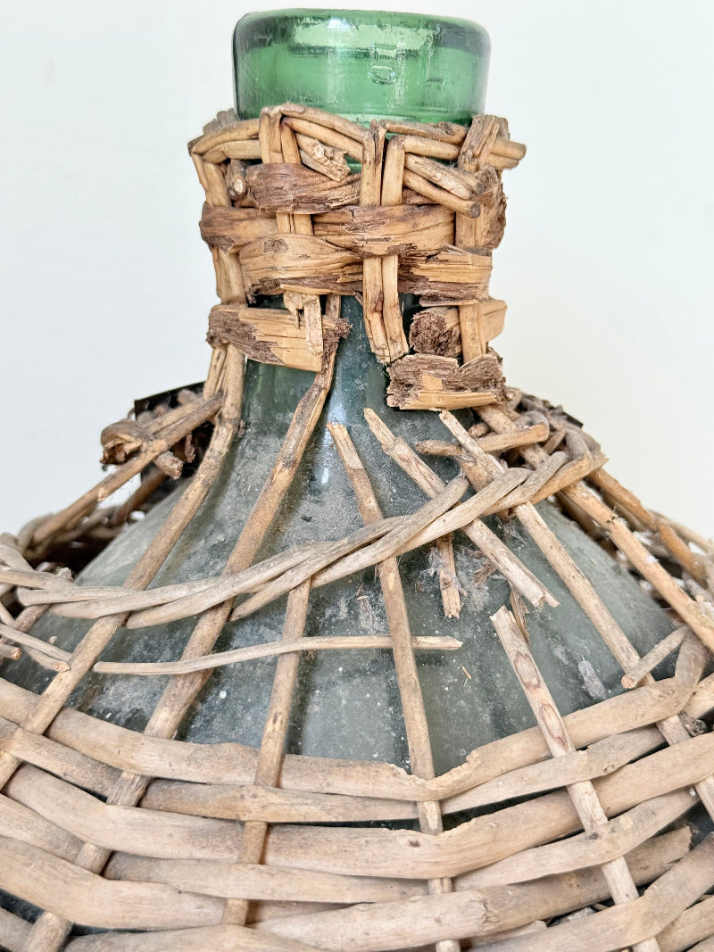 antique French demi john olive oil jar in wicker basket with green glass bottle and painted metal label, displayed on white surface