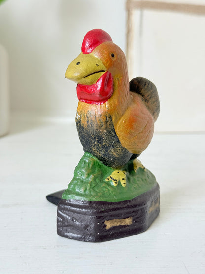 A hand-painted wooden rooster figurine standing on a green grassy hill atop a dark base, with a red comb and wattle, yellow beak, and black-and-brown feathered body.