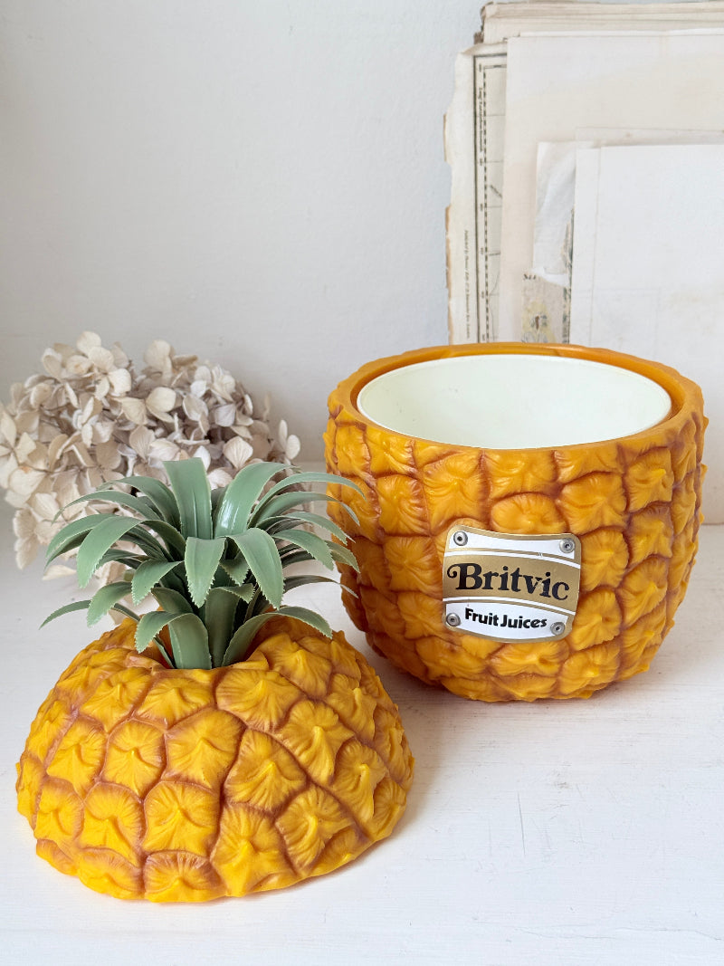 A novelty ceramic planter shaped like a pineapple, shown with its lid placed beside it on a white tabletop. The planter has a textured golden-yellow exterior, a green faux pineapple crown, and a silver metal label reading &