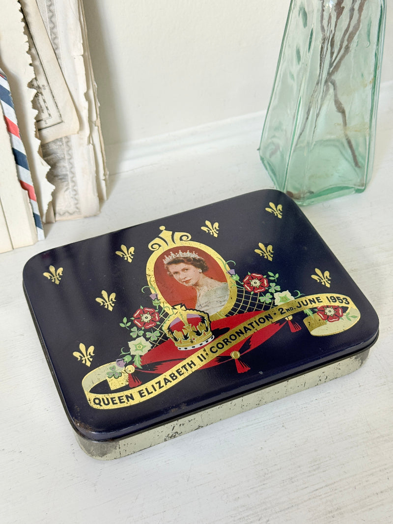 A square dark navy blue keepsake tin with a vintage Queen Elizabeth II Coronation design, resting on a white wooden surface next to a green glass vase.