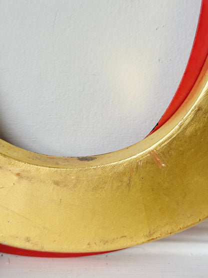 A close-up of a gold-colored circular frame or hoop with a red inner lining, shown against a plain off-white background.
