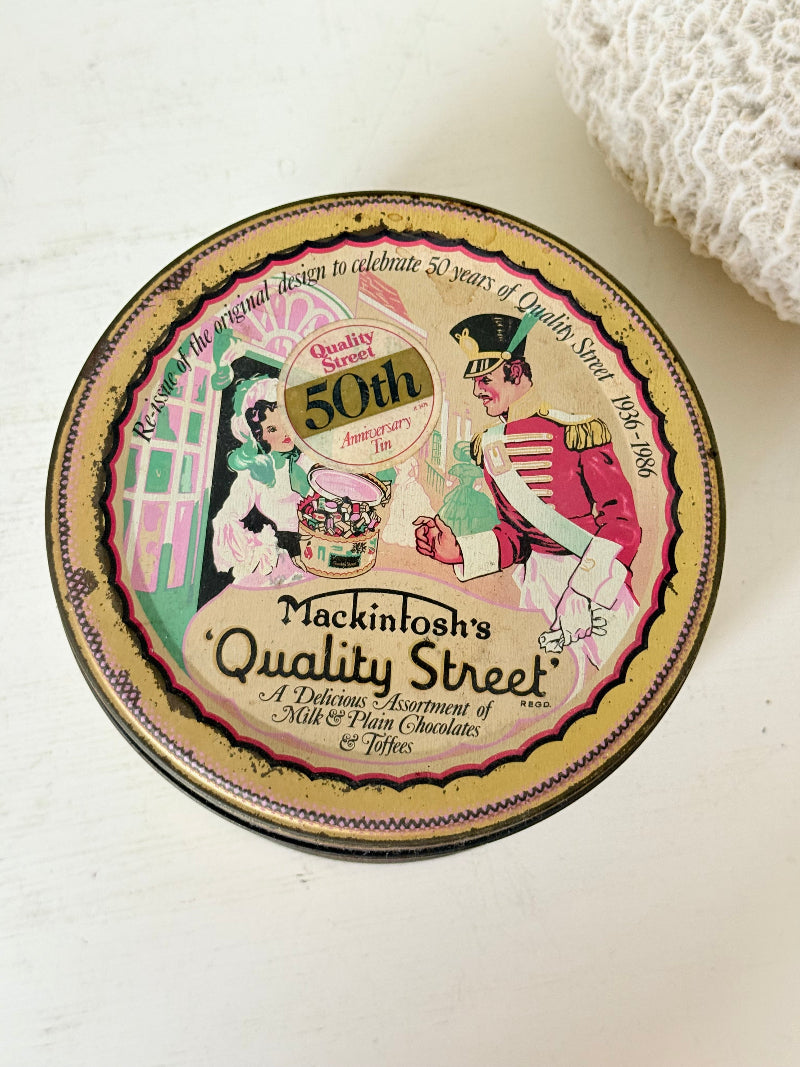 Vintage Quality Street tin from 1986 with colourful illustrated street scenes and pastel design, photographed on a white surface.