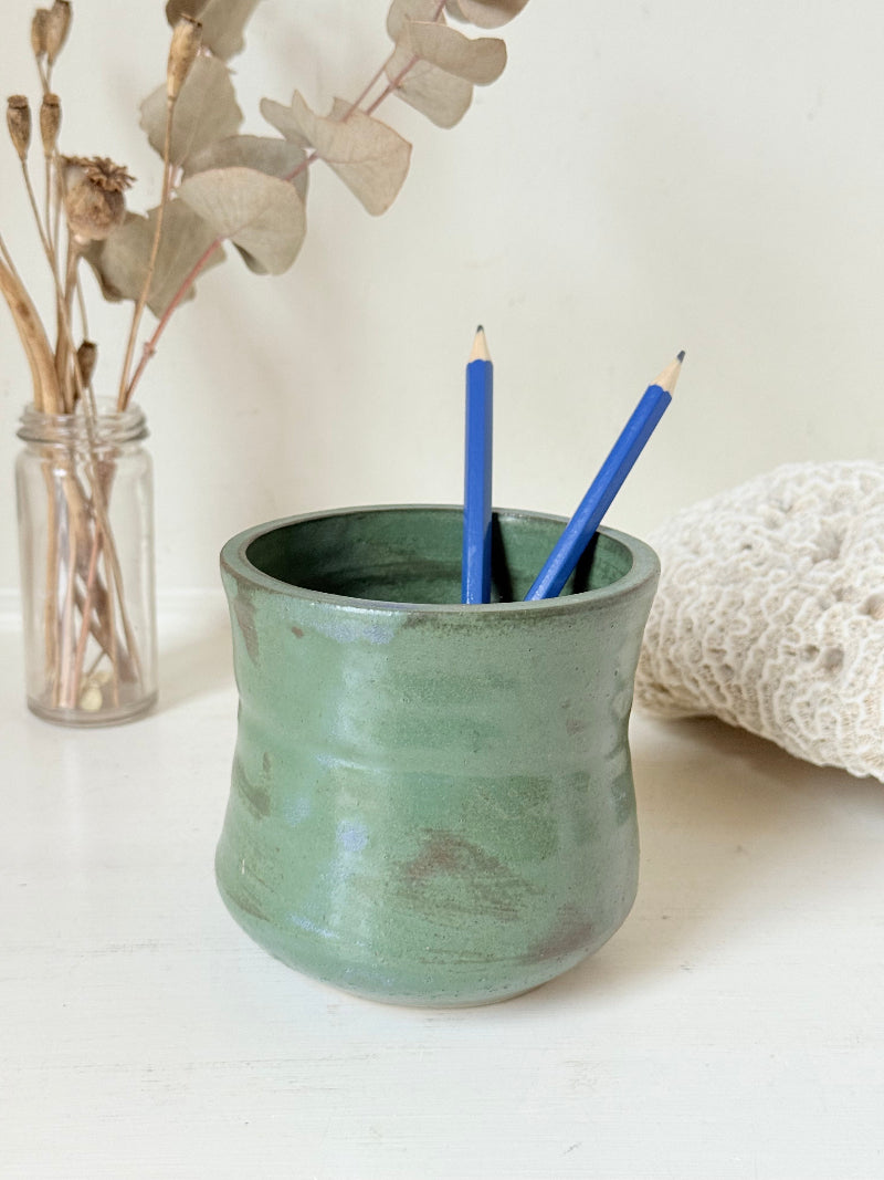 Studio pottery pencil pot in green glaze, handmade on the wheel, photographed on a white surface with pencils inside.