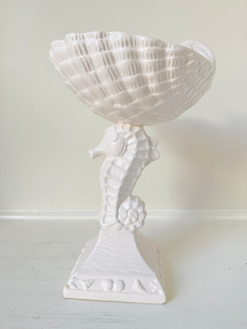 A white ceramic decorative bowl with a sculpted seahorse base, set against a plain off-white wall and surface.