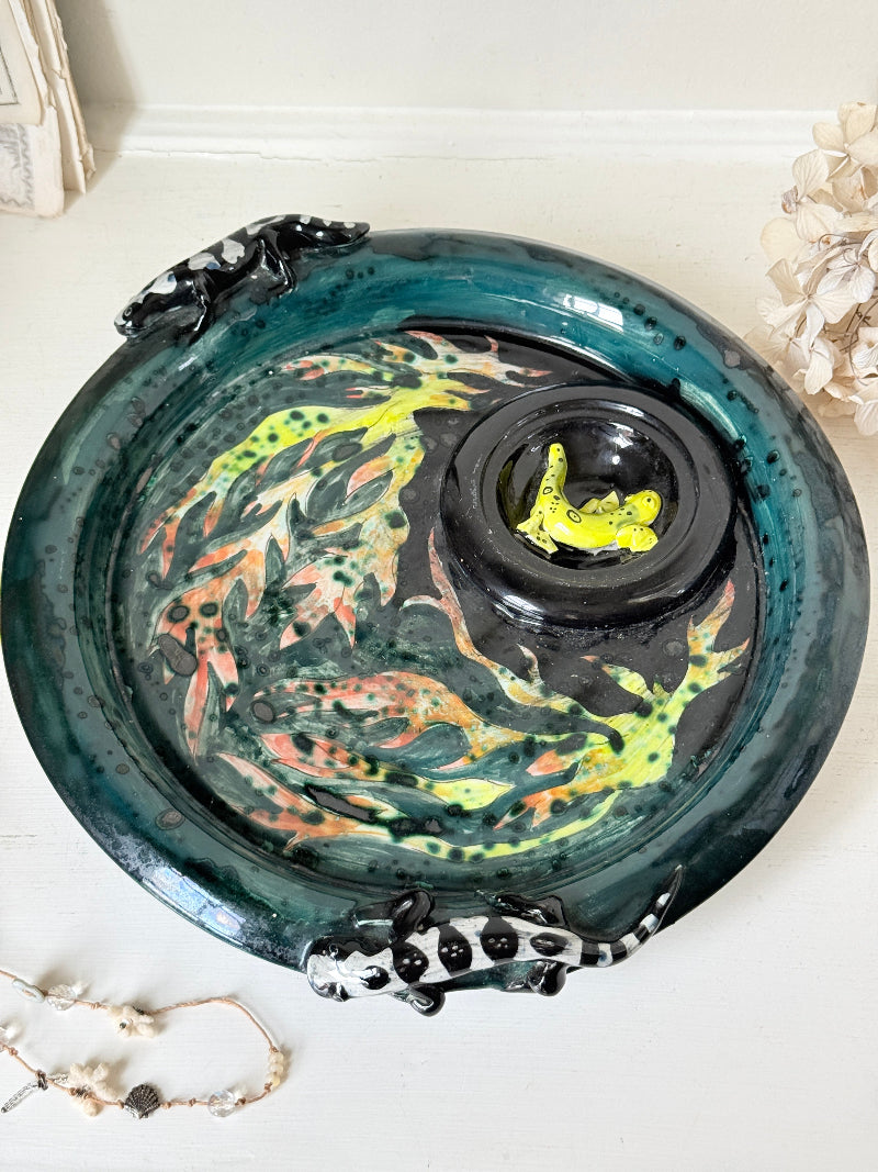 A decorative ceramic fish bowl with a glossy dark teal rim, hand-painted koi fish pattern in green, orange, and black, and a small black central pond section containing a bright yellow frog figurine. The bowl is styled on a white surface with dried flowers and a beaded necklace nearby.